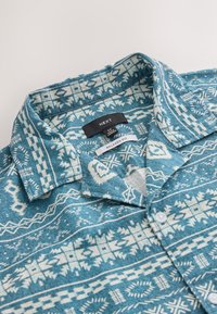 Kortærmet skjorte med løs pasform i teal med hvide stamme-mønstre. Stoffet er tekstureret; har krave og knaplukning foran. Brandetiket synlig.