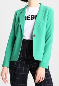 Blazer vert menthe avec une coupe ajustée et un col à revers. Présente un bouton unique et est porté par-dessus un t-shirt blanc et un pantalon à carreaux.