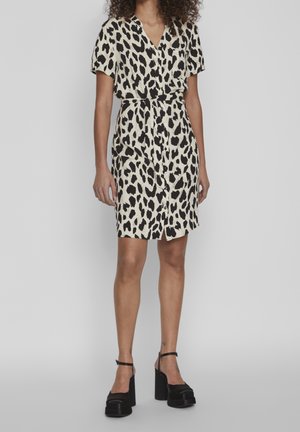 Femme portant une robe courte à manches courtes, longueur genou, avec un imprimé léopard noir et blanc, et des talons compensés noirs.