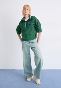Pull en polaire vert avec un col zippé à mi-hauteur, des accents marine et une poche. Un jean large bleu clair et des baskets crème complètent la tenue.