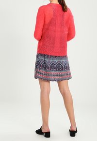 Korallenfarbener Strickpullover mit langen Ärmeln, strukturierter Stoff, kombiniert mit einem gemusterten Rock in Marineblau und Pink. Schwarze Schuhe mit Blockabsatz.