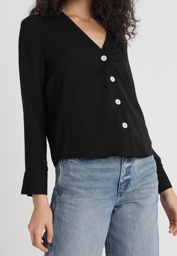 Femme portant un chemisier noir à col en V manches longues avec quatre boutons blancs et un jean taille haute bleu clair.