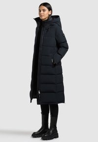 Abrigo acolchado largo negro con capucha, textura acolchada, cremallera frontal y bolsillos laterales. Combinado con botas negras hasta el tobillo y leggings.