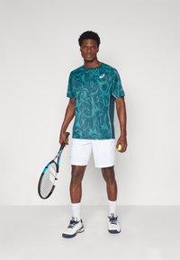 Türkiisin mustriga lühikeste varrukatega spordipop, mis on paaritatud valgete lühikesed pükstega. Mudel hoiab käes tennisereketit ja tennisepalli, kandes valgeid sokke ja spordijalatu. 