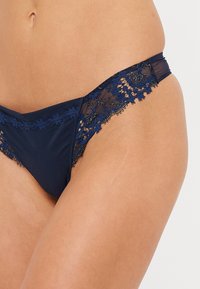 Jūras spēku zila bikini apakšbikses ar caurspīdīgu acs audumu un sarežģītu mežģīņu apdari ap jostu, ar gludu tekstūru un zemu vidukli.
