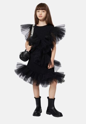 Vestido negro con volantes de tul negro en los hombros y en el dobladillo, combinado con botines negros hasta el tobillo. Acento con un pequeño bolso redondo.