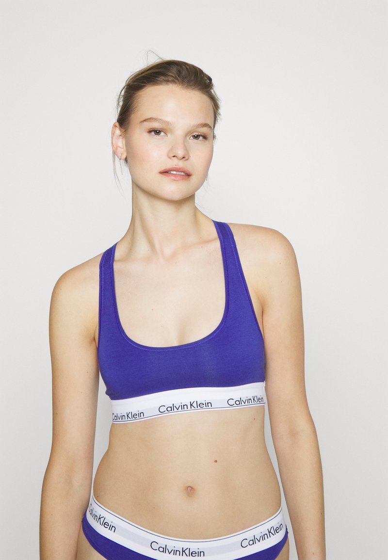 Fialová bavlnená športová podprsenka so širokým bielym elastickým pásom s logom "Calvin Klein". Jednoduchý výstrih na okrúhlo a ramienka v štýle tielka.