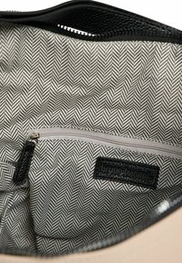 Schwarz-beige Handtasche mit Innenleben im Fischgrätmuster. Enthält ein Reißverschlussfach und ein schwarzes, strukturiertes Etikett. Insgesamt glatte Textur.