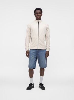 Mann steht, trägt eine leichte Jacke mit Reißverschluss, blaue knielange Jeansshorts, weiße Socken und schwarze Sneaker vor einfachem Hintergrund.