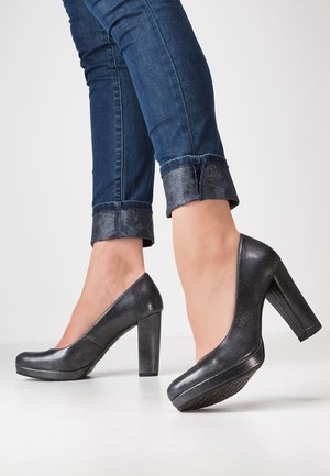 Person iført manchetterede mørkeblå jeans og sorte højhælede lædersko, stående på et lyst gulv foran en ensfarvet baggrund.