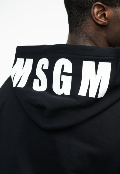 Gros plan sur le haut du dos d'une personne portant un sweat à capuche noir avec les grandes lettres blanches "MSGM" sur la capuche.