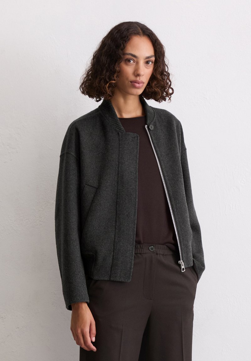 Donkergrijze wollen jas met een geribde kraag, voorkant met rits en zijzakken. Het ontwerp heeft een ontspannen pasvorm en lange mouwen.