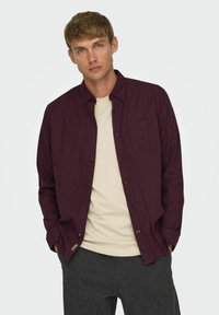 Bordeaux skjorte med knapper og lomme samt sorte knapper, båret over en beige t-shirt. Blød tekstur, stofblanding af bomuld. Casual pasform.