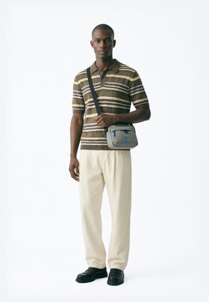 Mies, jolla on päällään raidallinen ruskea poolopaita, vaaleanbeiget housut, mustat kengät ja joka kantaa harmaata olkalaukkua sinisin yksityiskohdin, seisoo valkoista taustaa vasten.