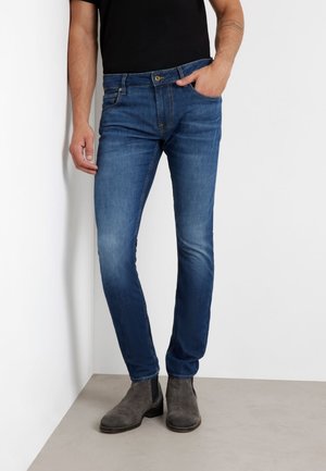 Jeans de mezclilla azul con un ajuste slim, diseño de cinco bolsillos y un ligero desgaste. Combinados con botas chelsea de ante grises.