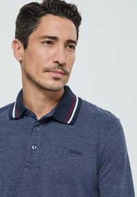 Homme avec des cheveux courts et foncés et une légère barbe, portant un polo bleu avec un col marine orné de rayures rouges et blanches.