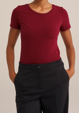 Kobieta w dopasowanej, krótkiej burgundowej koszulce, wsuniętej w wysokie czarne spodnie, z rękami w kieszeniach.