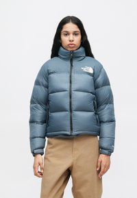 Osoba v modrej puffer jacket od North Face so zapnutým zipsom, stojí na jednofarebnom bielom pozadí s rukami pri bokoch.