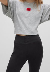 T-shirt gris court avec logo rouge "HUGO" et leggins taille haute noirs avec "HUGO" en relief sur la ceinture. Texture lisse, coupe décontractée.