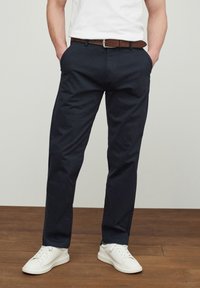 Chinos marinos de algodón con un diseño de pierna recta, que cuentan con bolsillos laterales y un cinturón de cuero marrón. Combinados con zapatillas blancas.