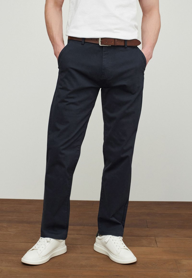 Chinos marinos de algodón con un diseño de pierna recta, que cuentan con bolsillos laterales y un cinturón de cuero marrón. Combinados con zapatillas blancas.
