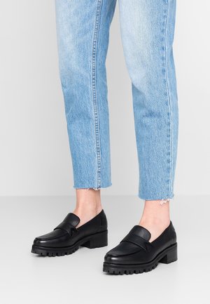 Sorte læder loafers med en kraftig sål og en tekstureret overdel, prydet med et dekorativt bånd over vristen. Båret med lyseblå jeans.