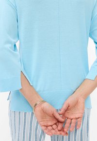 Lichtblauwe gebreide trui met driekwartmouwen, zijsplitten en een ronde hals, gecombineerd met gestreepte broek en gouden armbanden.