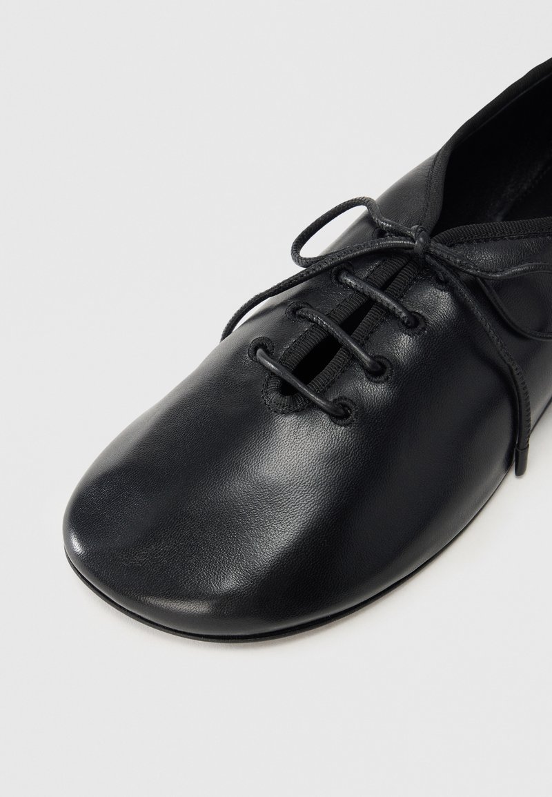 Chaussure en cuir noir à bout arrondi, avec un design à lacets et des accents élastiques. Texture lisse et style minimaliste.