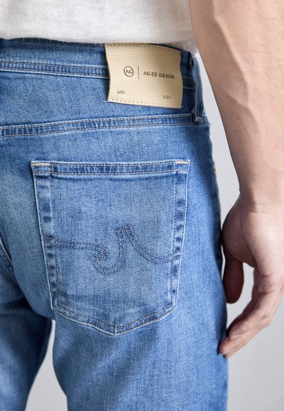 Svetlomodré džínsové nohavice s textúrovanou koženou nášivkou, s prehnutým dizajnom zadnej vrecka a viditeľnými švami.