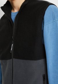 Svart och grå fleeceväst med hög krage, zippered front och subtil branding. Den nedre delen har en slät textur.