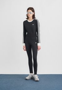 Žena oblečená v čiernom Adidas teplej obleku s bielymi pruhmi, šedých športových sandáloch, bielych ponožkách a odrazových slnečných okuliaroch, stojaca na modrom koberci.