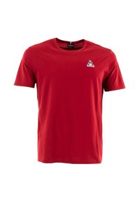 Camiseta de algodón roja con mangas cortas y cuello redondo. Presenta un pequeño logotipo azul y blanco en la parte izquierda del pecho. Textura suave.