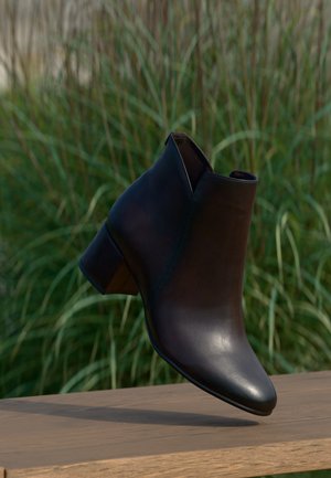 Bottine noire en cuir avec un petit talon bloc, exposée au-dessus d'une surface en bois, herbe verte en arrière-plan.