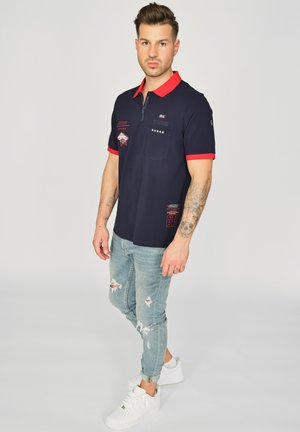 Marine polo med rød krave, lynlås og frontlomme; har hvid syning og dekorative lapper. Bæres med lyse, slidte jeans og sneakers.