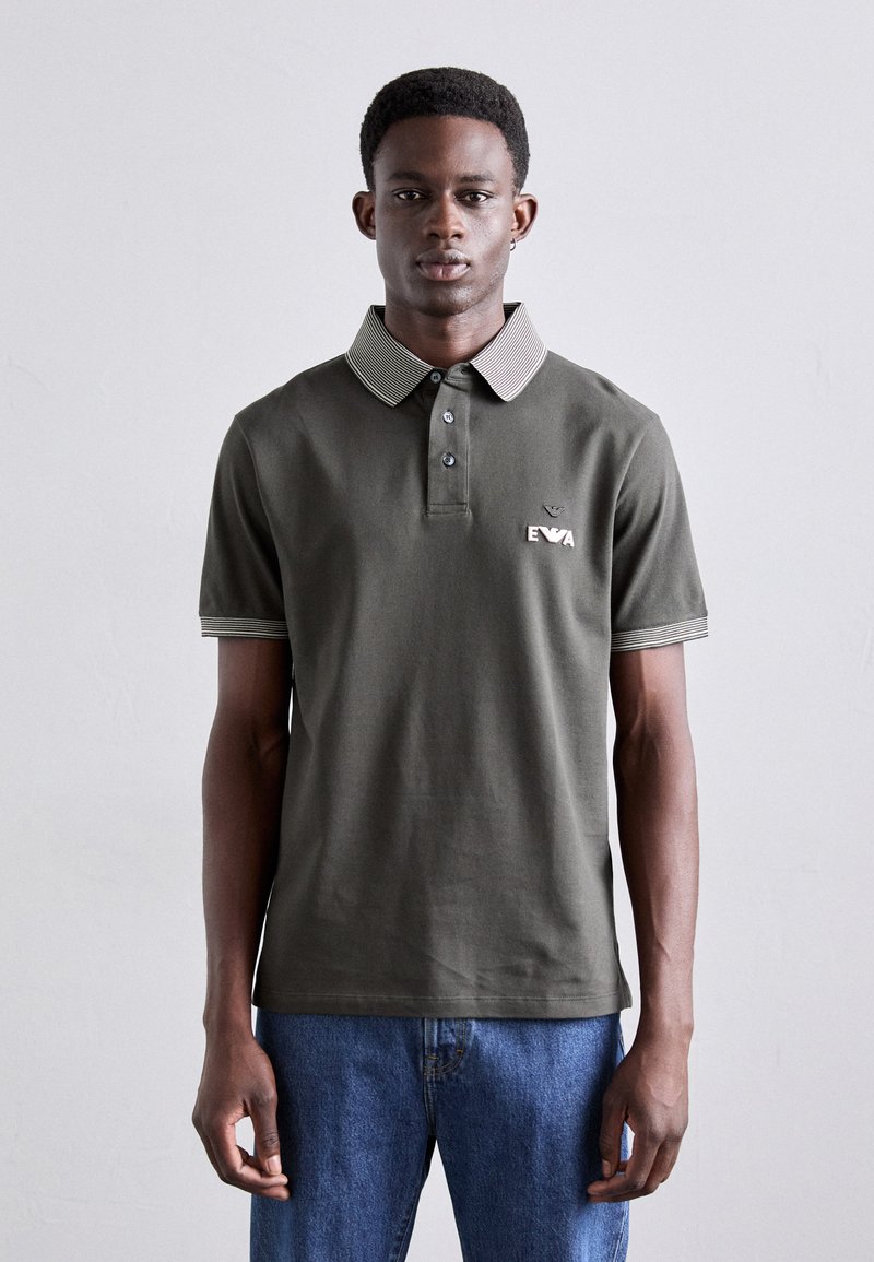 Tumši zaļš polo krekls, izgatavots no kokvilnas, ar svītrotu apkakli un piedurkņu malām, trīs pogu aizdari un izšūtu logotipu uz krūtīm.