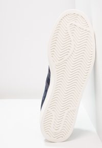 Tmavě modrý semišový teniska s texturovanou gumovou podrážkou s vlnitým vzorem a měkkou bílou střední podrážkou, nakloněná na jednoduchém pozadí.