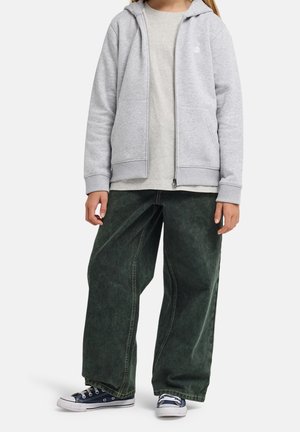 Kind draagt een lichtgrijze hoodie met rits, beige shirt, brede donkergroene broek en marineblauwe sneakers op een witte achtergrond.