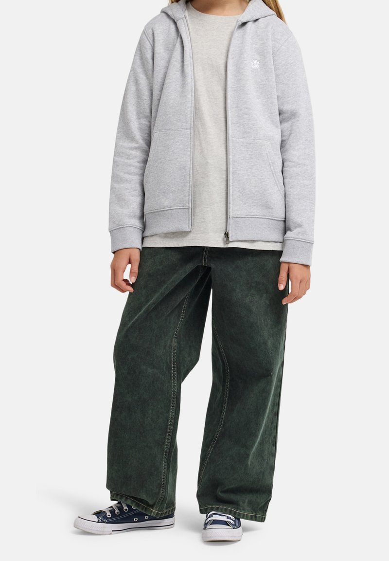 Dieťa oblečené v svetlošedej mikine na zips, béžovom tričku, širokých tmavozelených nohaviciach a námorníckych modrých teniskách na bielom pozadí.