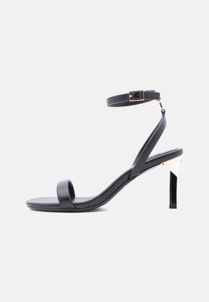 Sort hæl-sandal med stropper, ankelspænde, slank hæl og åben tå mod en ensartet hvid baggrund.
