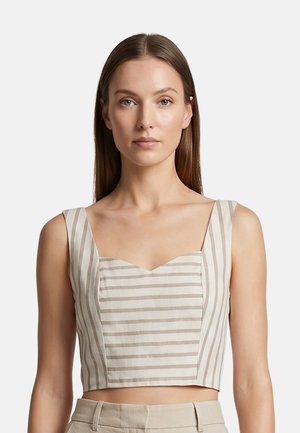 Vrouw met steil bruin haar, gekleed in een beige-wit gestreept mouwloos cropped topje en beige broek tegen een effen witte achtergrond.