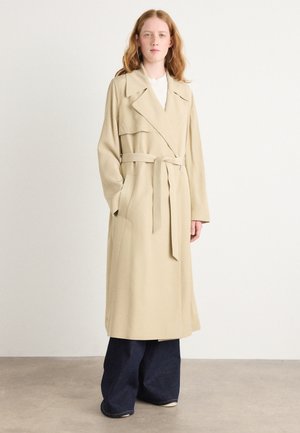 Manteau beige avec une taille ceinturée, des revers larges et des manches longues. Présente une texture lisse et tombe au niveau du mollet. Jeans noirs à larges jambes en dessous.