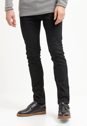 Person trägt schlanke schwarze Hose, graues Langarmoberteil und schwarze Lederschuhe mit braunen Sohlen und steht vor weißem Hintergrund.