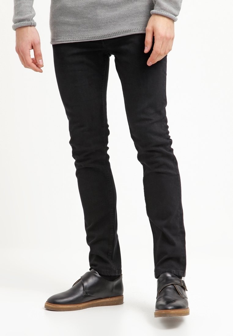 Personne portant un pantalon noir slim, un haut à manches longues gris et des chaussures en cuir noir avec des semelles marron, debout devant un fond blanc.