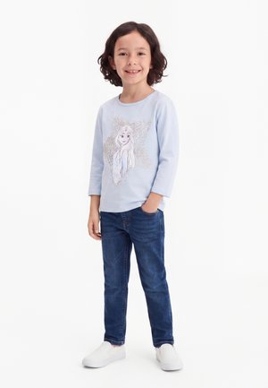 Bambino sorridente con capelli ricci che indossa una camicia a maniche lunghe azzurra con un personaggio animato, jeans blu e scarpe bianche senza lacci, in piedi con la mano in tasca.