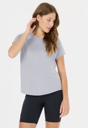 Kvinde med langt brunt hår iført en lysegrå t-shirt og sorte, tætsiddende shorts, stående med den ene hånd berørende sit hår.