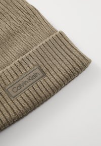 Khaki gebreide beanie met geribbelde textuur, voorzien van een rechthoekig bruin label met de opdruk "Calvin Klein" op de boord.