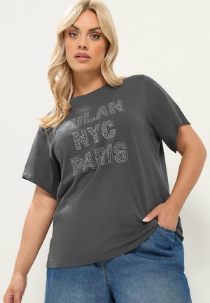 Blondynka w ciemnoszarej koszulce z napisem "MILAN NYC PARIS" i niebieskich dżinsach, stojąca z jedną ręką w kieszeni na neutralnym tle.