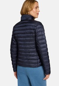 Veste matelassée bleu marine avec un design matelassé, un col montant et une finition brillante. Présente une silhouette ajustée et des poignets élastiques pour la chaleur.