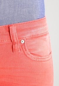 Acercamiento de un bolsillo de mezclilla color coral y un lazo del cinturón, usados con una camisa azul clara metida por dentro.