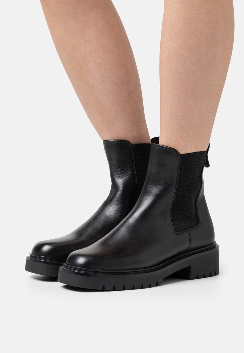Bottines en cuir noir lisse avec semelles épaisses portées sur jambes nues sur un fond clair uni.